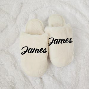 Cream-colored slippers personalized with name “James,” displayed on soft white blanket — cozy men’s gift.