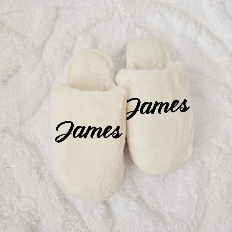 Cream-colored slippers personalized with name “James,” displayed on soft white blanket — cozy men’s gift.
