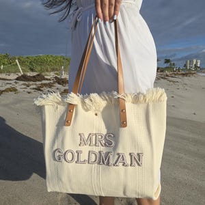 Close-up of bride in white dress holding ivory “Mrs Goldman” personalized beach tote with pearl letters and woven fabric texture.