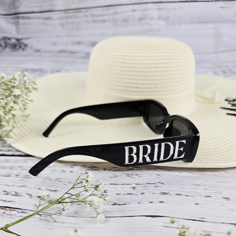 Black BRIDE sunglasses on ivory “Mrs.” hat with white florals on a light wood background, perfect for bridal photo styling.