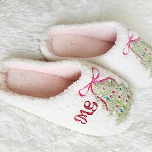 May include: A pair of white, plush slippers with pink interiors. Each slipper features an embroidered Christmas tree design with colorful accents and the word "me" in red glitter.