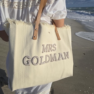 Bride holding ivory “Mrs Goldman” personalized tote bag with pearl letters and tan leather handles near ocean waves.