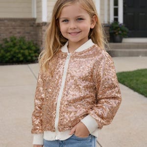 May include: A rose gold sequin bomber jacket with a white zipper and white ribbed cuffs and collar. The jacket is worn with blue jeans.
