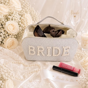 May include: A silver cosmetic bag with the word "BRIDE" spelled out in pearl-like beads. The bag is open, revealing sunglasses and a hair scrunchie. A champagne flute with "Bride" printed on it and a candle are also visible.