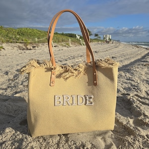 Beige bridal tote bag with pearl “BRIDE” lettering and fringe top detail placed on sandy beach with ocean and palm trees in background.