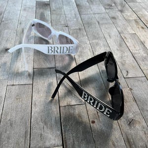 White and black BRIDE sunglasses on wooden flooring, minimal aesthetic perfect for bridal flat-lay photography.
