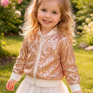 May include: A young girl smiles, wearing a gold sequin bomber jacket with white trim and a white tulle skirt. The jacket has a zipper closure. The background features green grass and pink flowers.