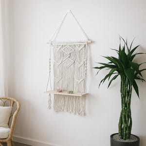 May include: Cream-colored macrame wall hanging with a wooden shelf. The macrame features a geometric diamond pattern and fringe. Two small decorative items rest on the shelf. A tall green plant in a black pot is to the right.