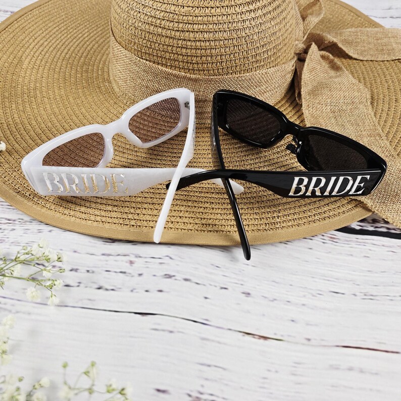 Stylish black and white BRIDE sunglasses placed on a tan straw hat, ideal bridal accessories for beach or honeymoon photos.