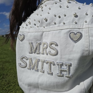 May include: White denim jacket embellished with pearls and rhinestones. The back features the name "MRS SMITH" in pearl lettering, with heart-shaped patches. The jacket is decorated with scattered pearls and rhinestones.