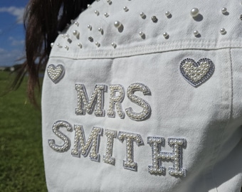 May include: White denim jacket embellished with pearls and rhinestones. The back features the name "MRS SMITH" in pearl lettering, with heart-shaped patches. The jacket is decorated with scattered pearls and rhinestones.
