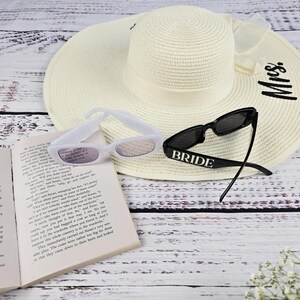 Flat lay of black and white BRIDE sunglasses with sun hat and open book, creating a chic summer bridal aesthetic.