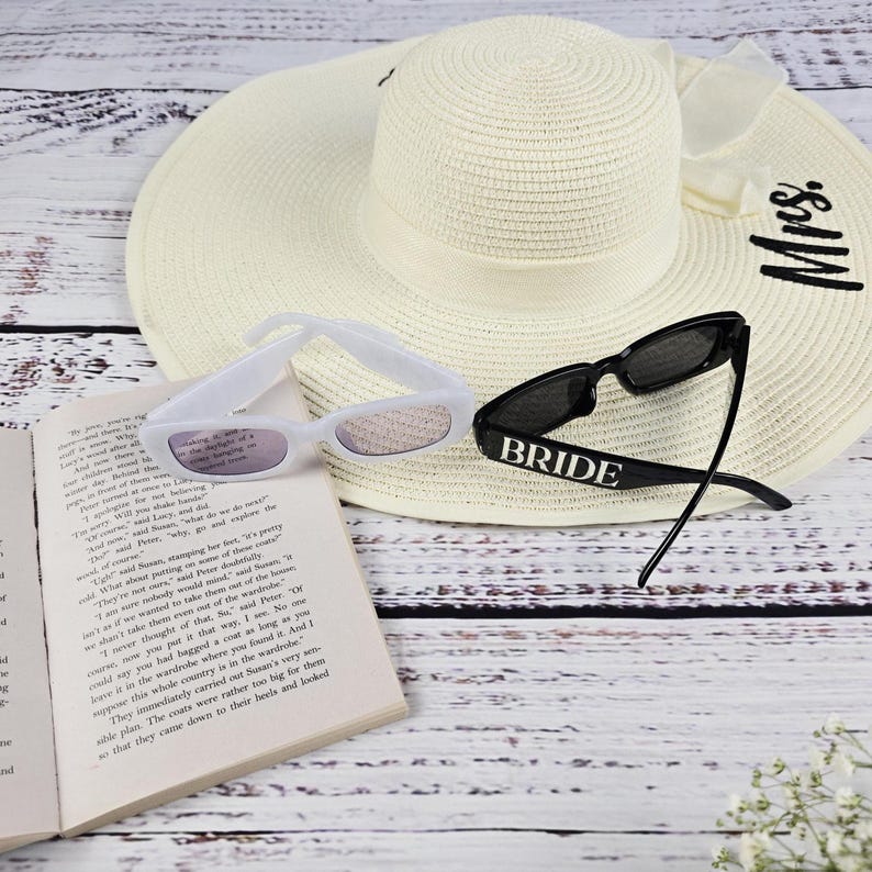 Flat lay of black and white BRIDE sunglasses with sun hat and open book, creating a chic summer bridal aesthetic.