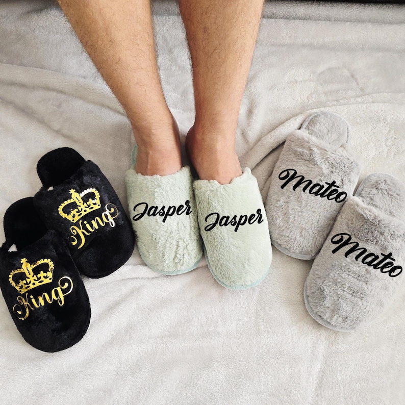 Man wearing mint green slippers with the name Jasper, surrounded by black “King” slippers and grey “Mateo” slippers — personalized men's footwear.