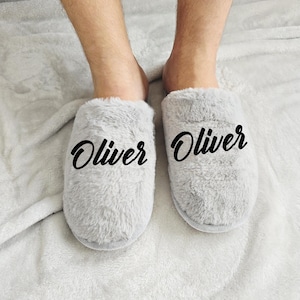 Close-up of grey plush slippers with the name Oliver written in black lettering, modeled on soft blanket background.