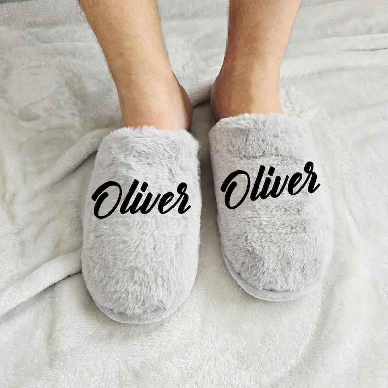 Close-up of grey plush slippers with the name Oliver written in black lettering, modeled on soft blanket background.