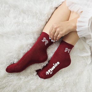 May include: Burgundy-colored socks with a white bow design near the ankle. One sock has the name "Monica" printed in white script. The socks are ribbed at the top and are displayed on a white, fluffy surface.