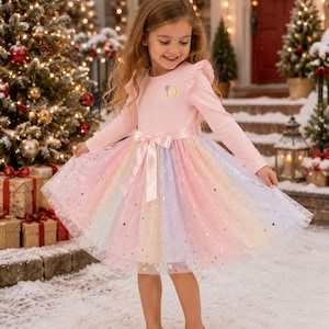 May include: A long-sleeved dress with a light pink ribbed bodice featuring a small letter D embroidered on the chest. The skirt is made of multi-colored pastel tulle layers adorned with small silver stars and a satin ribbon bow at the waist.