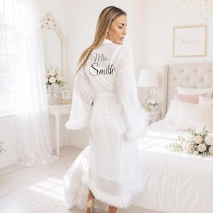 May include: A sheer white robe featuring long, flowing sleeves and a hemline trimmed with white feathers. The back of the robe displays the text "Mrs. Smith" in a black, elegant script font. The garment includes a matching tie belt at the waist.