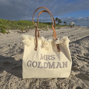 Ivory “Mrs Goldman” bridal beach bag with pearl letters and fringe top displayed on sand with palm trees in the background.