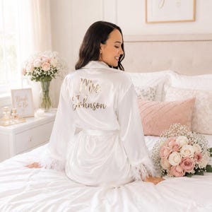 May include: A white satin robe with feather trim, featuring gold text that reads "Mrs. Johnson." The robe is tied at the waist and worn by a person sitting on a bed. A bouquet of roses and baby's breath is nearby.