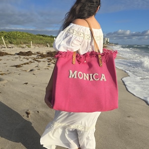 Bride in white dress holding pink personalized tote with pearl “Monica” letters and fringe top by the ocean.