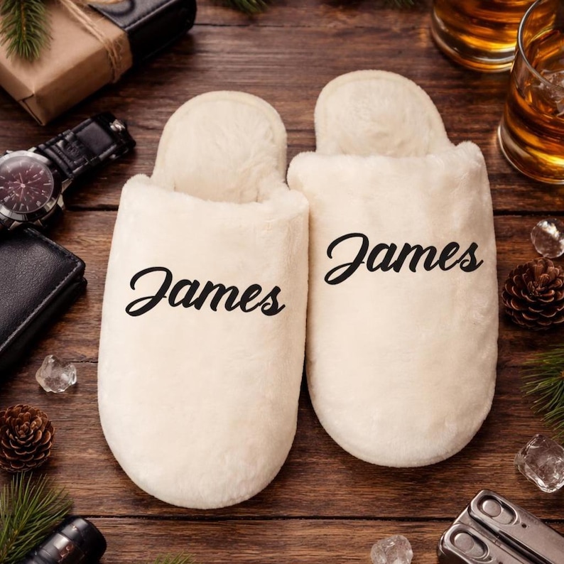 May include: A pair of cream-colored, plush slippers with the name "James" printed in black script on each slipper. The slippers are on a wooden surface, with other items like a watch and a wallet in the background.
