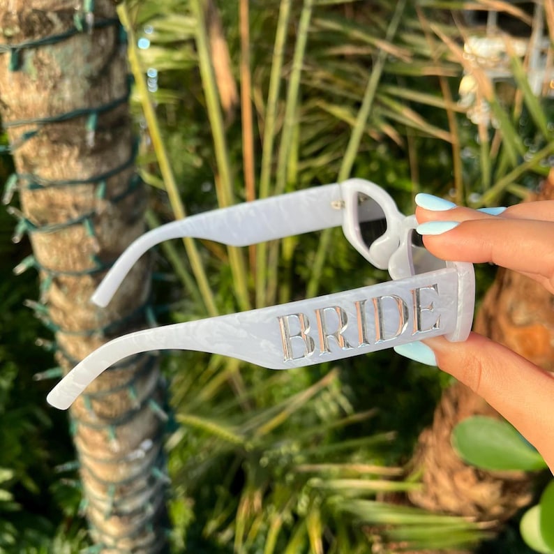 White BRIDE sunglasses held outdoors with palm trees in the background, showing reflective metallic lettering under natural sunlight.