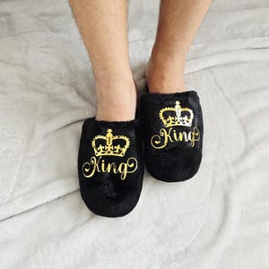 Man modeling black plush “King” crown slippers with gold vinyl design — perfect gift for husband or boyfriend.