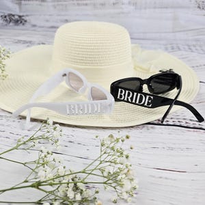 White and black BRIDE sunglasses displayed on a white summer hat with delicate white flowers, perfect for beach weddings or bachelorette parties.