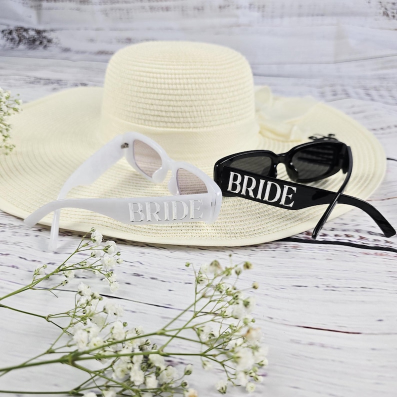 White and black BRIDE sunglasses displayed on a white summer hat with delicate white flowers, perfect for beach weddings or bachelorette parties.