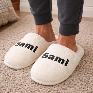 May include: A pair of white, fluffy slippers with the word "Sami" printed in black on each slipper. The slippers are on a beige carpet. The person wearing the slippers is wearing gray sweatpants.