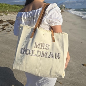 Bride carrying ivory woven tote bag with pearl “Mrs Goldman” letters and tan leather handles while walking along a sunny beach shoreline.