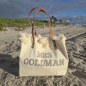 Ivory woven bridal tote “Mrs Goldman” with pearl lettering photographed on beach sand under golden sunlight.