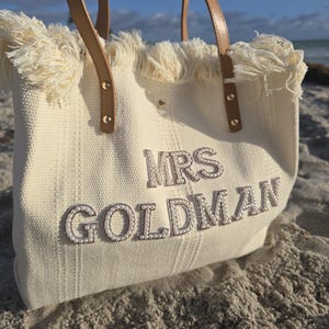 Detailed beach close-up of ivory bridal tote “Mrs Goldman” showing pearl letters, fringe detail, and woven fabric texture.