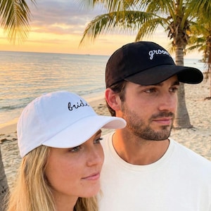 May include: A white baseball cap with the word "bride" embroidered in black script and a black baseball cap with the word "groom" embroidered in white script. The caps are worn by a couple on a beach.