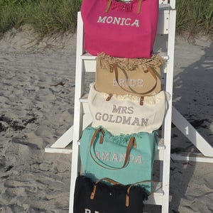 Group of four custom bridal tote bags stacked on white chair — “Monica,” “Bride,” “Mrs Goldman,” “Amanda,” and “Rachel” — in pink, beige, ivory, mint, and black colors.