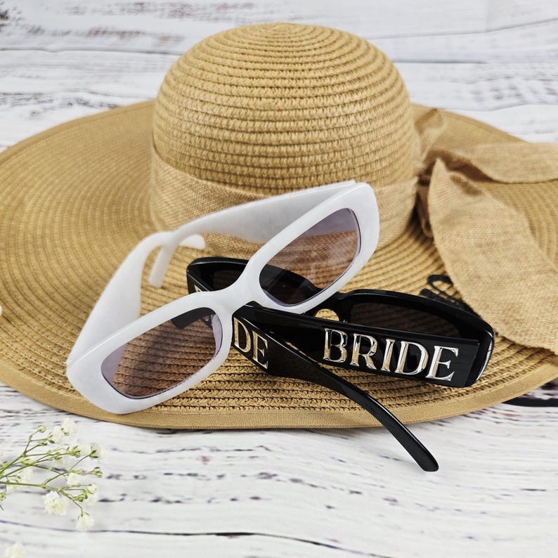 White and black BRIDE sunglasses on a tan straw hat with rustic wood background, styled for bridal beachwear photography.