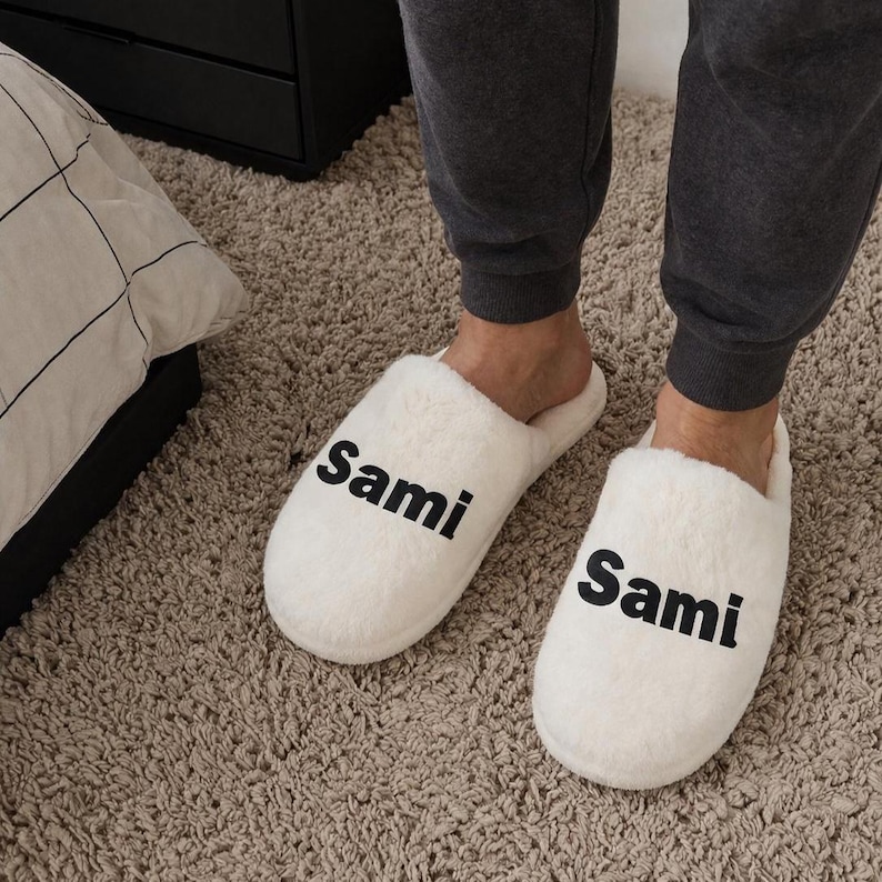 May include: A pair of white, fluffy slippers with the name "Sami" printed in black on each slipper. The slippers are on a beige carpet.