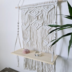 May include: A macrame wall shelf with a natural wood shelf and intricate woven design. The shelf is suspended by a rope and holds a pink decorative item and a silver bracelet. The macrame is white.