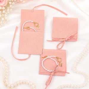 May include: A collection of pink suede jewelry pouches and pearl bracelets. The bracelets feature gold clasps and initial charms. One pouch is open, revealing a bracelet with the letter "L". Another bracelet has the letter "H". The pouches are tied with pink ribbons.