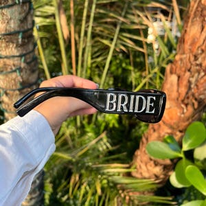 Black BRIDE sunglasses held outdoors with palm trees and sunlight reflection, showing shiny embossed letters.