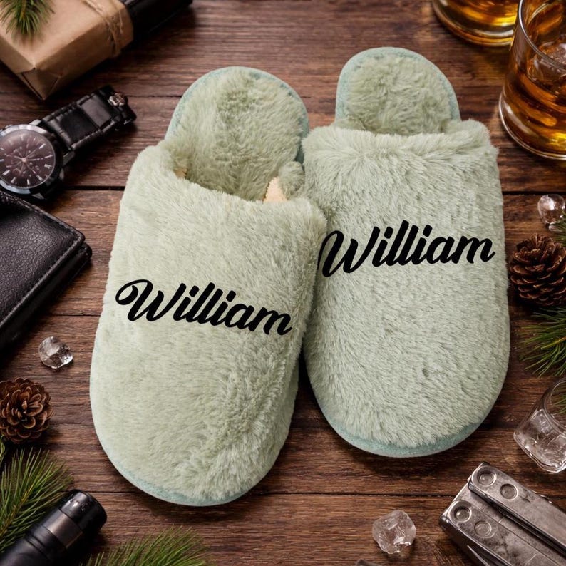 May include: A pair of light green plush slippers with the name "William" printed in black script. The slippers are on a wooden surface, with a watch, wallet, and other items in the background.