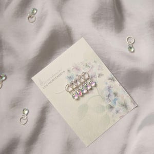 Può includere: Orecchini color argento con ciondoli iridescenti a forma di goccia sono presentati su una carta con illustrazioni floreali e la scritta "Les Hortensias". Ulteriori orecchini sono sparsi su uno sfondo di tessuto bianco.