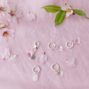 May include: A collection of delicate jewelry charms and connectors, featuring silver-toned rings and clear or pink crystal beads. The charms are arranged on a soft pink surface, adorned with scattered flower petals and blossoms.