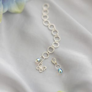 Peut inclure: Un bracelet à chaîne argentée composé d'une série de maillons circulaires texturés, d'une breloque papillon en filigrane délicat et d'une petite perle en forme de goutte irisée. Un fermoir mousqueton argenté avec une perle assortie est inclus.