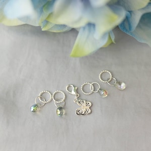 May include: Silver-toned stitch markers with iridescent crystal beads and a butterfly charm. The stitch markers are arranged on a gray surface, with a blurred blue and yellow flower in the background. These are ideal for knitting and crochet projects.