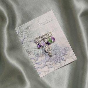 May include: A silver-toned brooch featuring a lavender bouquet charm and dangling gemstone beads in purple and green hues. The brooch is displayed on a card with the text "La Lavanderie Les Lavandes" and a lavender illustration.