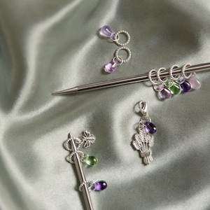 Könnte beinhalten: Eine Sammlung von Strickmaschenmarkierern und Zubehör. Die Marker haben silberne Ringe mit lila, grünen und klaren tropfenförmigen Perlen. Ein Marker hat einen Weizen-Anhänger und eine lila Perle. Die Artikel liegen auf einem Satin-Hintergrund.