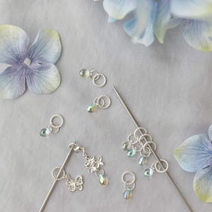 May include: A collection of silver hair accessories and decorative elements, including hair sticks, rings, and a butterfly charm, are arranged on a light gray surface. Blue and purple hydrangea flowers are scattered around the accessories.